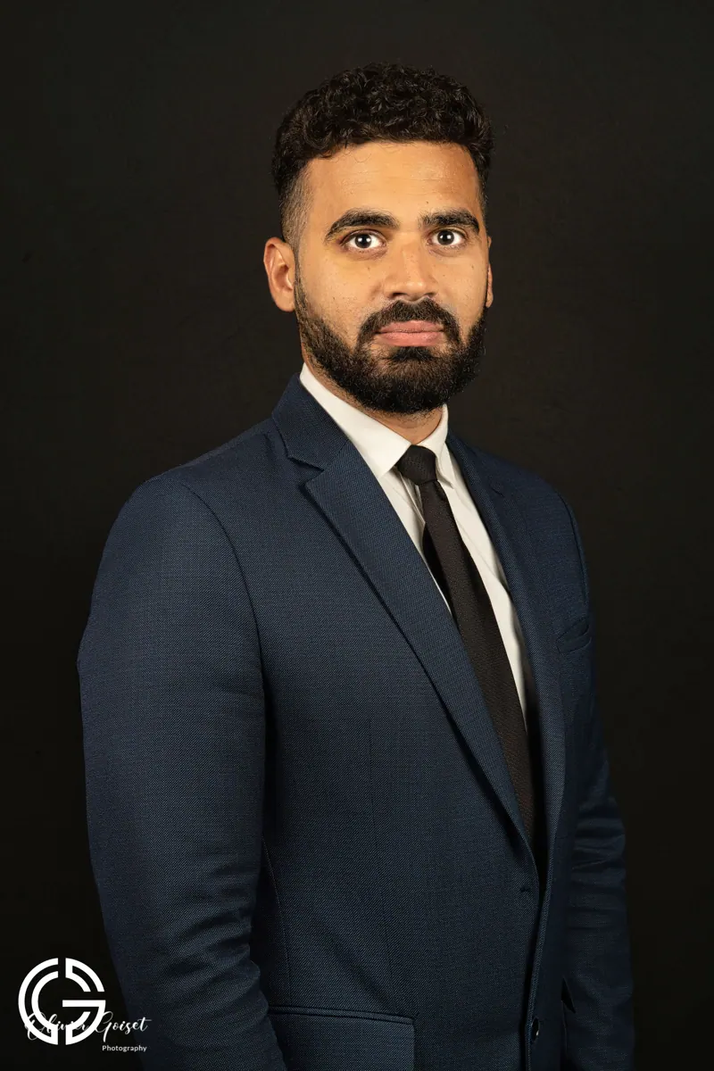 Portrait d'un homme barbu en costume bleu et cravate noire, regard droit, sur fond noir.