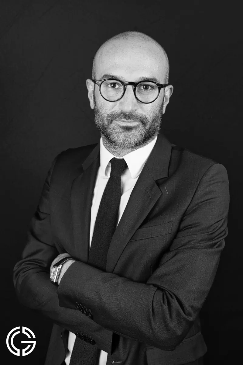 Portrait en noir et blanc d'un homme à lunettes, costume sombre et cravate, bras croisés.