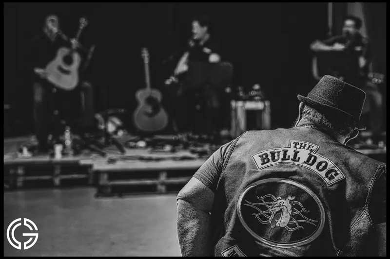 De dos, un homme au blouson brodé « The Bull Dog » regarde une scène de concert, en noir et blanc.