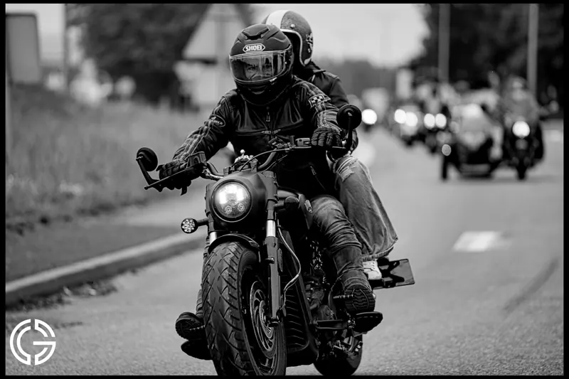 Un motard et sa passagère roulent en tête d'un convoi, en noir et blanc.