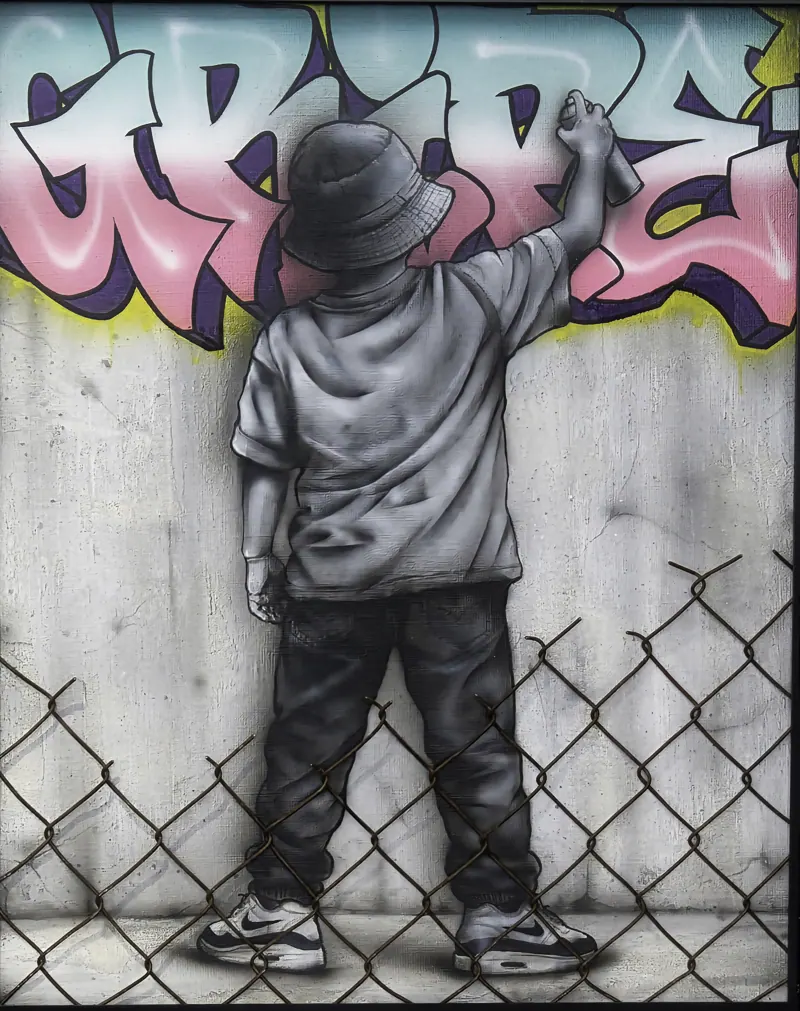 Un enfant en casquette tague un mur derrière un grillage.